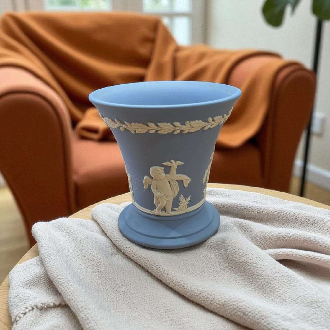Jarrón Wedgwood azul jaspeado con relieve estilo clásico único en mesa de madera, decorando un espacio acogedor con sillón naranja.