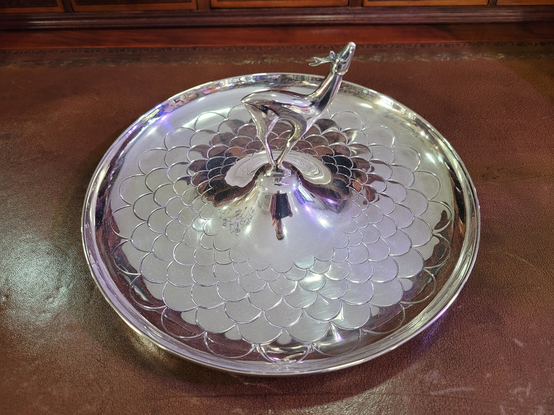 Cenicero de metal plateado con detalle de ciervo en relieve, diseño elegante y decorativo sobre fondo de cuero marrón.