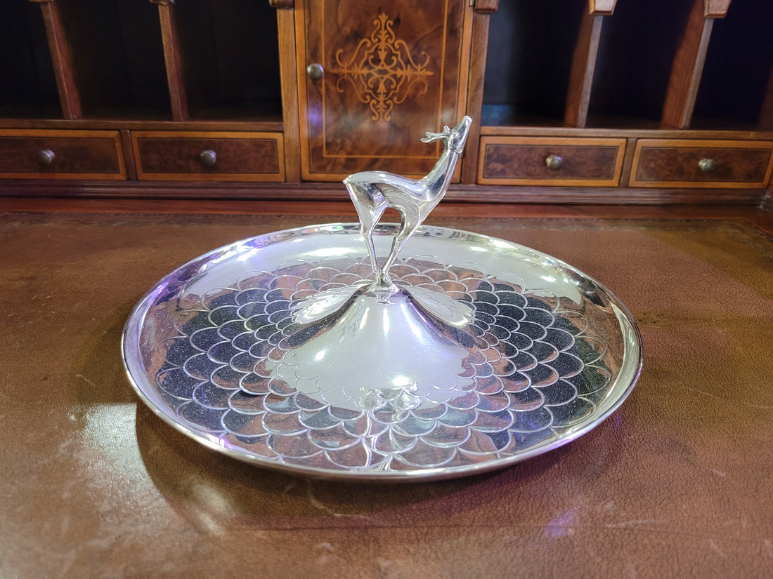 Cenicero de metal plateado con detalle de ciervo en relieve, diseño elegante y decorativo sobre fondo de cuero marrón.