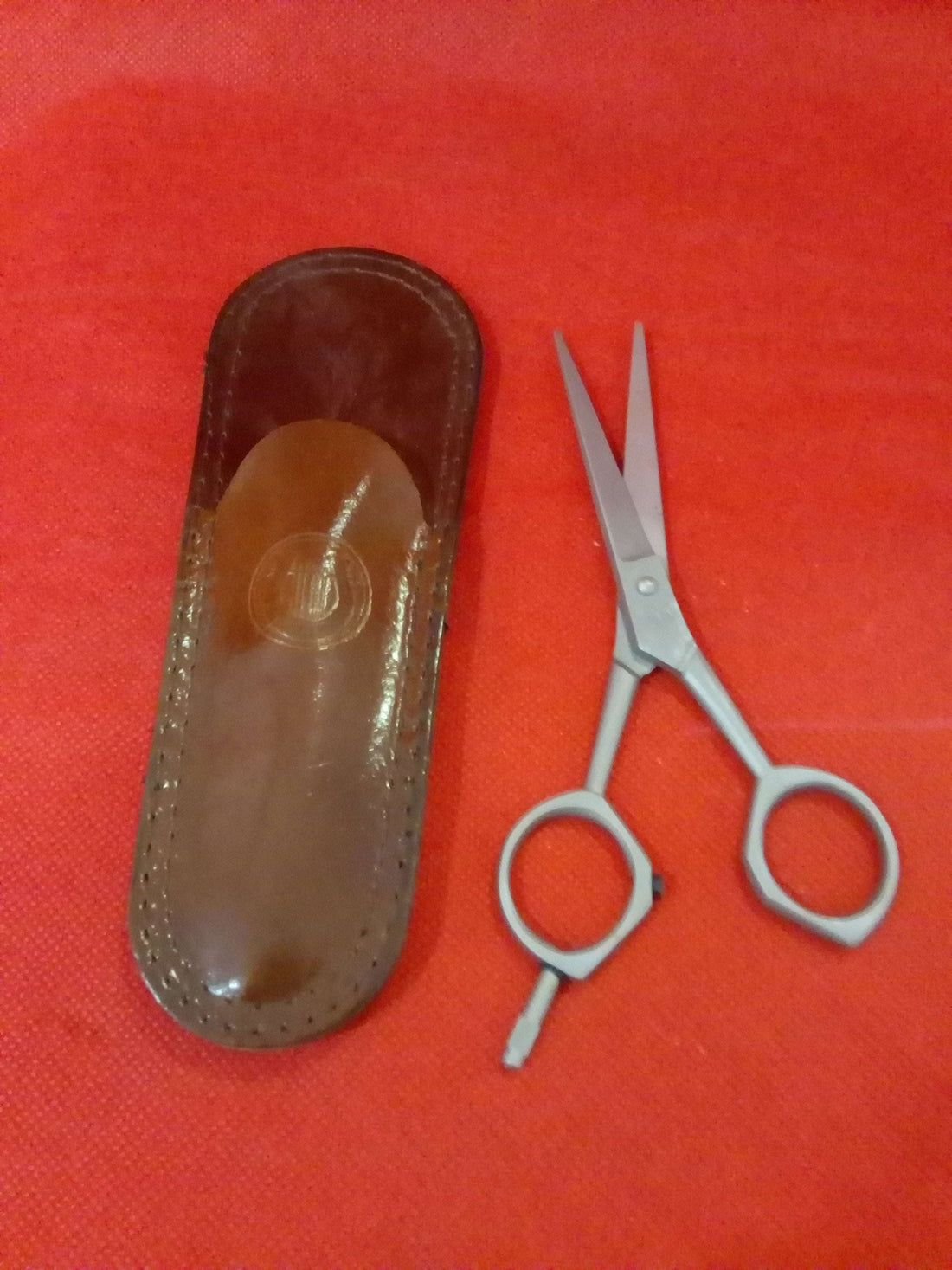 Tijera de barbero Filarmónica de cobalto con diseño vintage junto a su funda original de cuero sobre fondo rojo. Ideal para decoración temática.