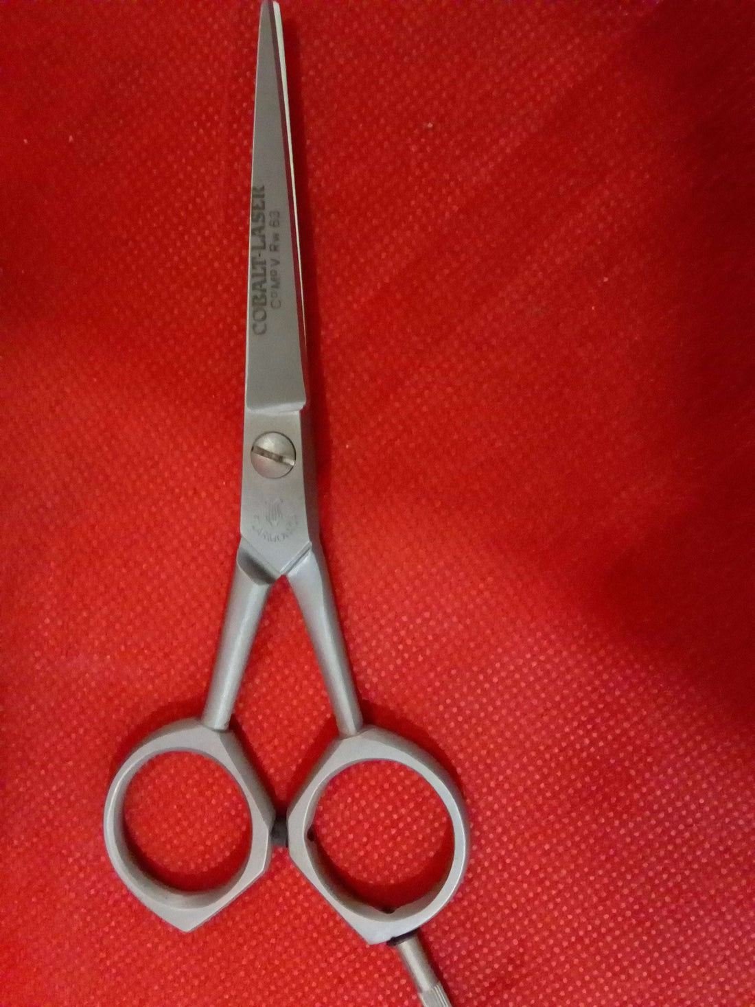 Tijera de barbero Filarmónica de cobalto con diseño vintage junto a su funda original de cuero sobre fondo rojo. Ideal para decoración temática.