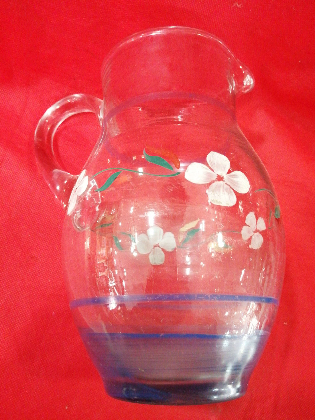 Jarra de cristal antigua decorada a mano con flores blancas y hojas verdes sobre fondo rojo. Ideal para decoración vintage en el hogar.