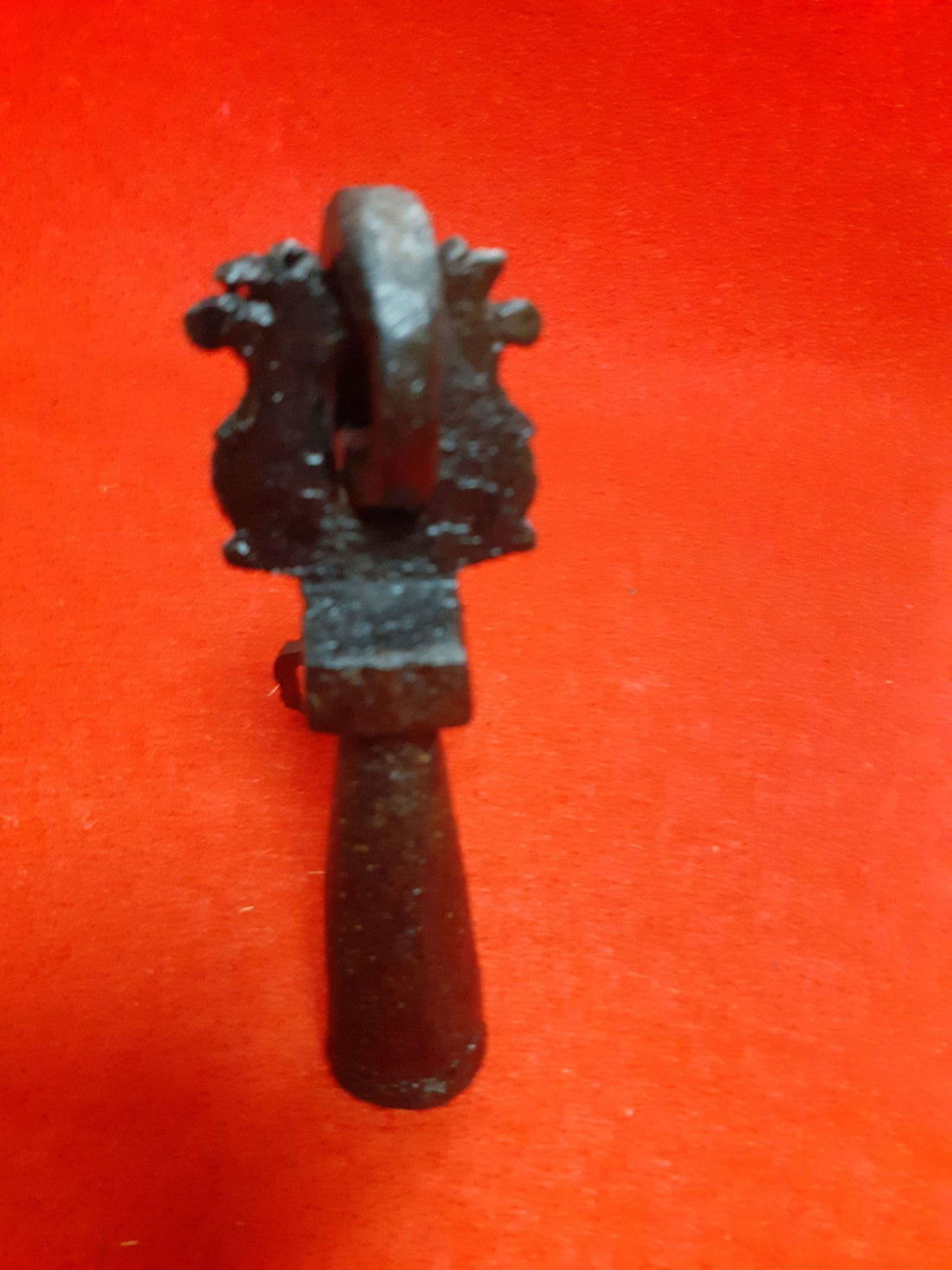 Aldaba-llamador de puerta antiguo S. XVIII en hierro forjado con diseño ornamental sobre fondo rojo, ideal para decoración vintage en el hogar.