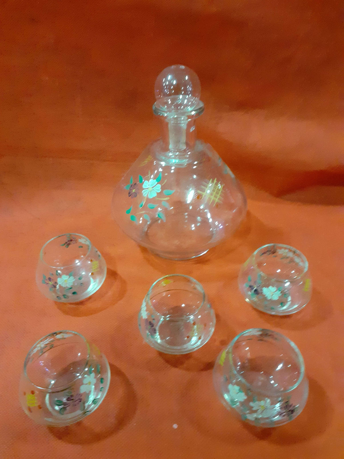 Botella licorera pintada a mano con 5 vasitos de cristal sobre fondo naranja. Diseño floral ideal para decoración de hogar y ocasiones especiales.