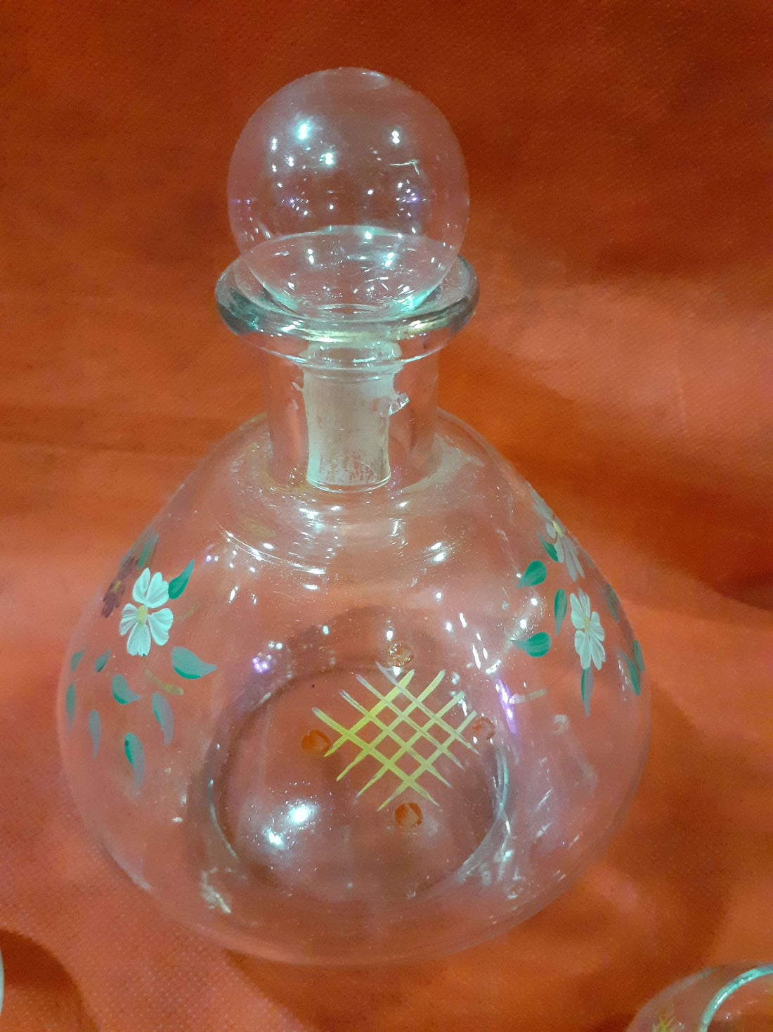 Botella licorera pintada a mano con 5 vasitos de cristal sobre fondo naranja. Diseño floral ideal para decoración de hogar y ocasiones especiales.