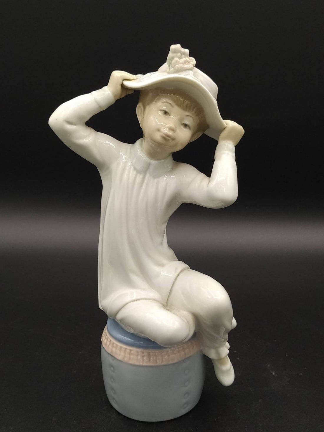 Figura de porcelana Lladró 'Chica con gorro' sentada sobre un tambor azul, con vestido blanco y sombrero decorado, ideal para decoración elegante.