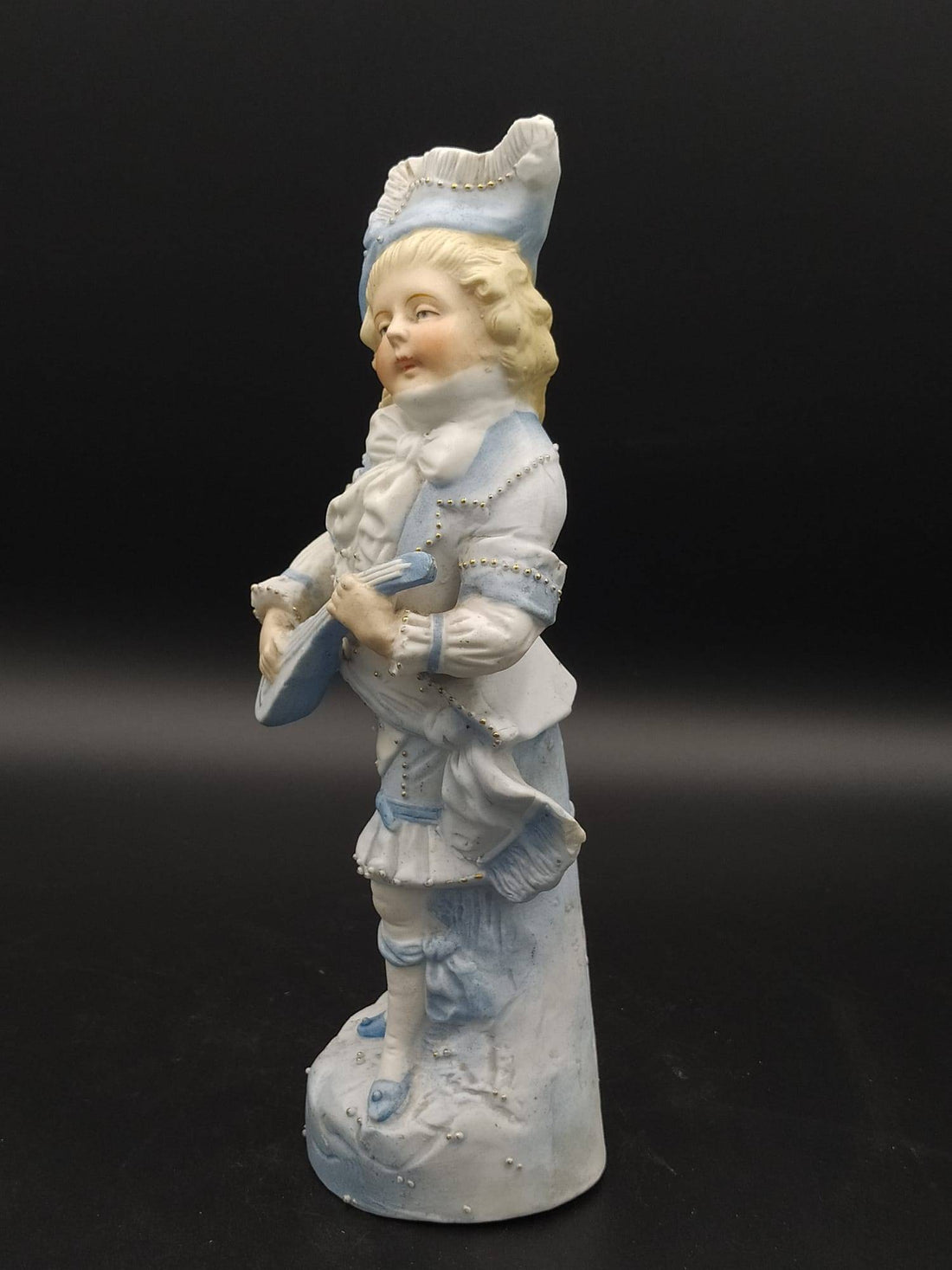 Figura de porcelana antigua de galleta, niño tocando cuerda, excelente, con vestimenta azul estilo rococó sobre fondo negro.