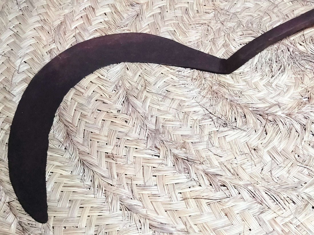 Hocino de brazo largo y curvado con mango de madera sobre fondo de palma tejida. Decoración rústica para jardín o pared interior.