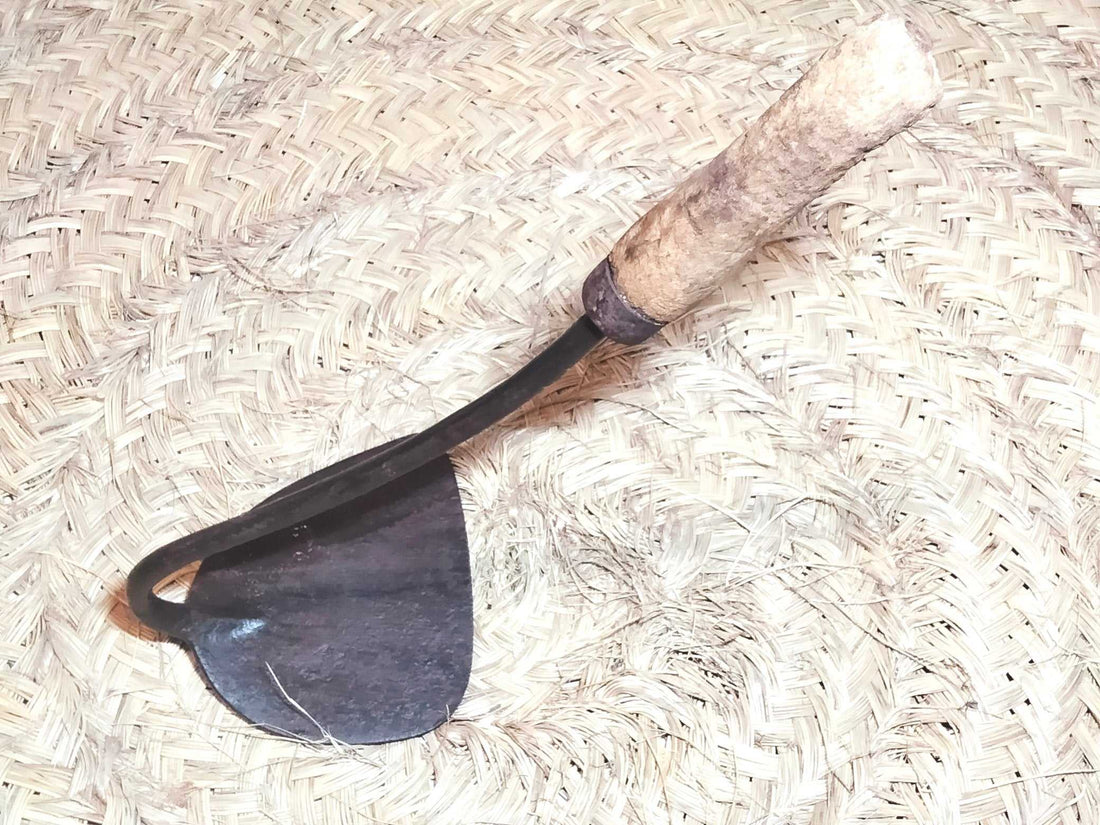 Amocafre con mango de madera curvo sobre fondo de palma tejida. Herramienta de jardín única ideal para decoración rústica o uso práctico.