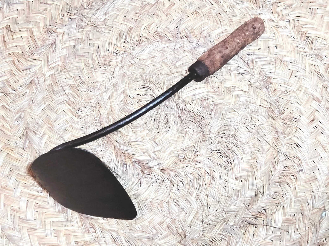 Amocafre con mango de madera curvo sobre fondo de palma tejida. Herramienta de jardín única ideal para decoración rústica o uso práctico.