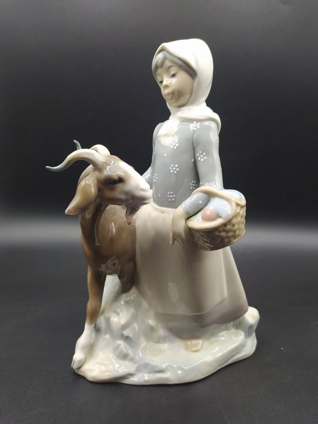 Figura Lladró 'Niña con cabra' en porcelana, muestra a una niña con pañuelo y canasta junto a una cabra. Decoración clásica de excelente calidad.