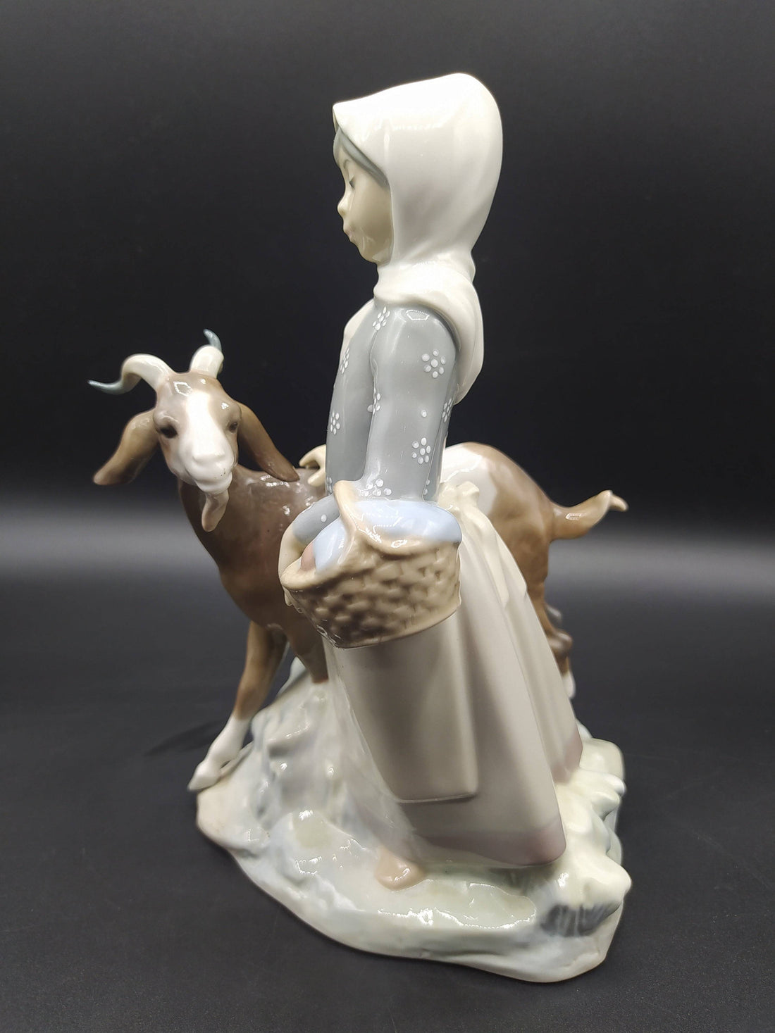 Figura Lladró 'Niña con cabra' en porcelana, muestra a una niña con pañuelo y canasta junto a una cabra. Decoración clásica de excelente calidad.