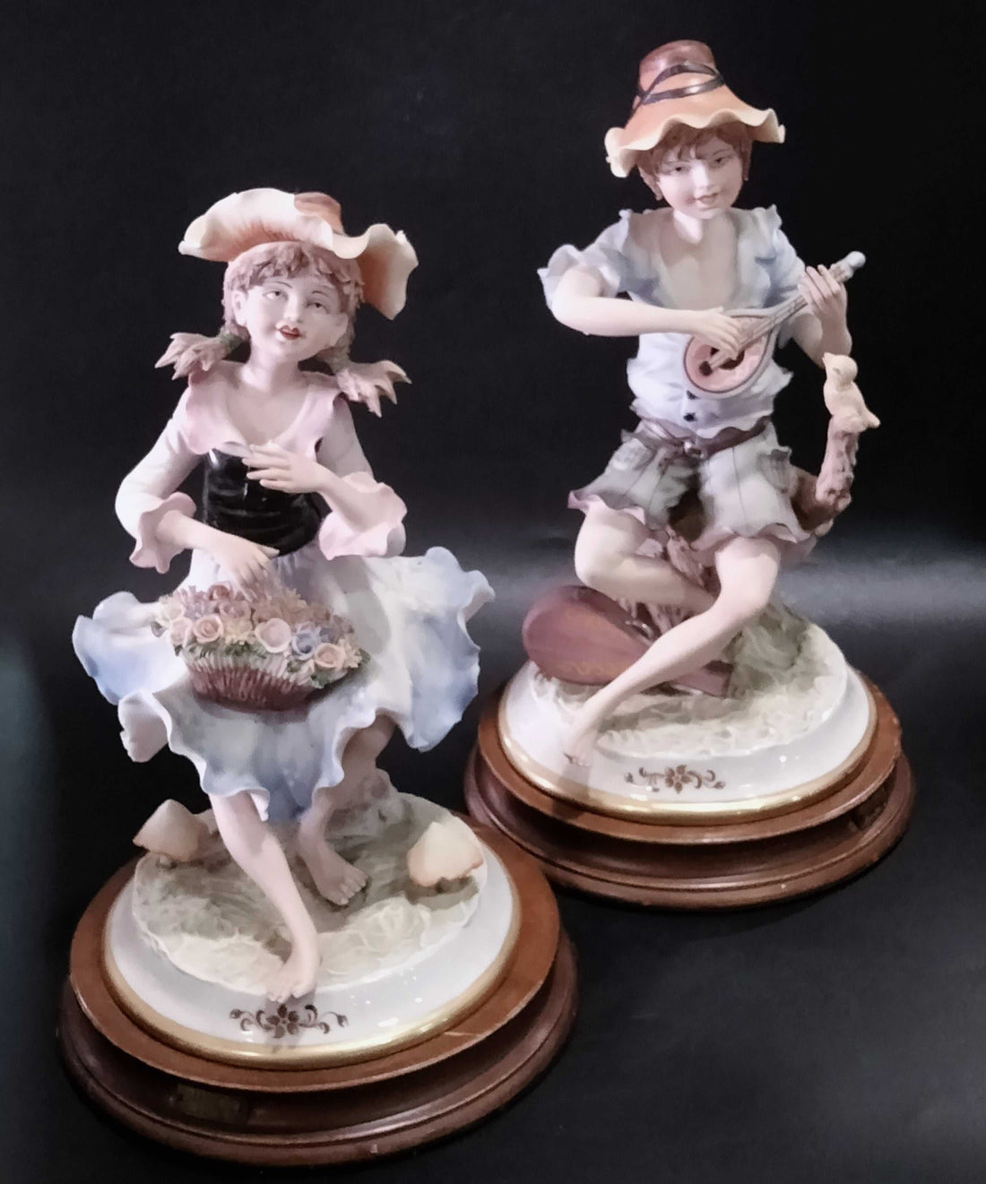 Figuras de porcelana fina marca Carlos | Niño con instrumento y niña con flores sobre metopas de madera, decoración elegante para el hogar.