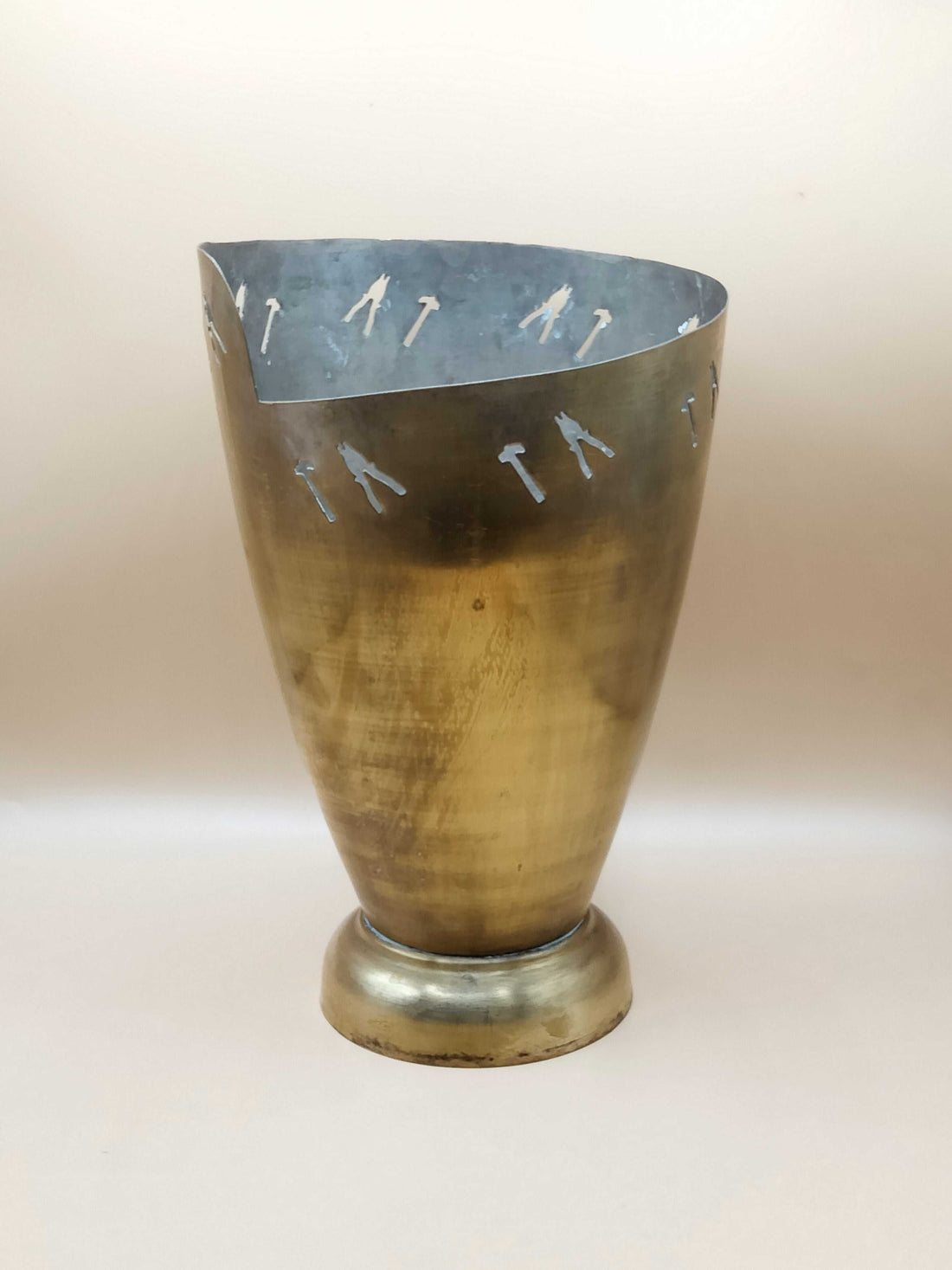 Jarrón de metal con detalles artesanales, único en su estilo, acabado dorado y figuras recortadas de herramientas en la parte superior.