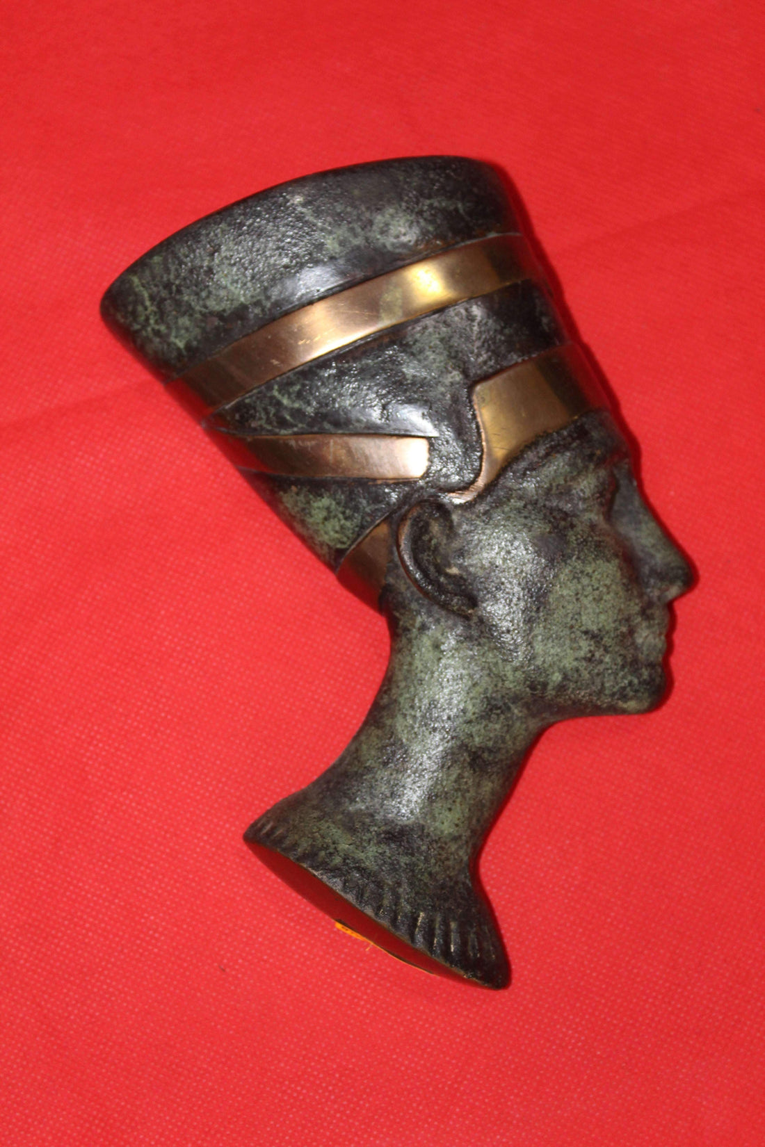 Busto de Nefertiti en metal con acabado envejecido y detalles dorados sobre fondo rojo. Réplica antigua ideal para decoración y coleccionistas.