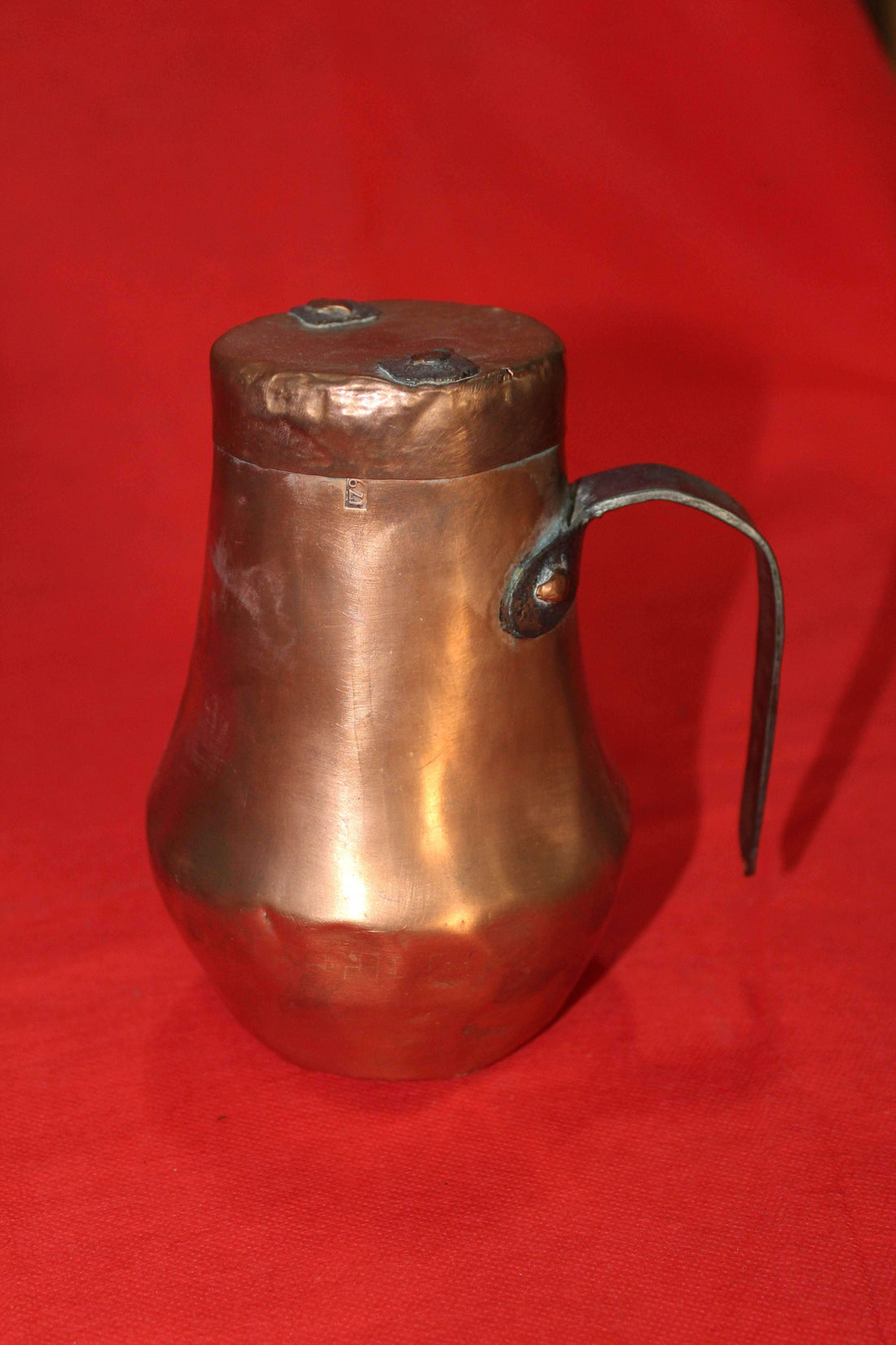 Jarra antigua de cobre con tapadera sobre fondo rojo, ideal para decoración vintage en hogar y jardín. Diseño rústico con asa metálica artesanal.