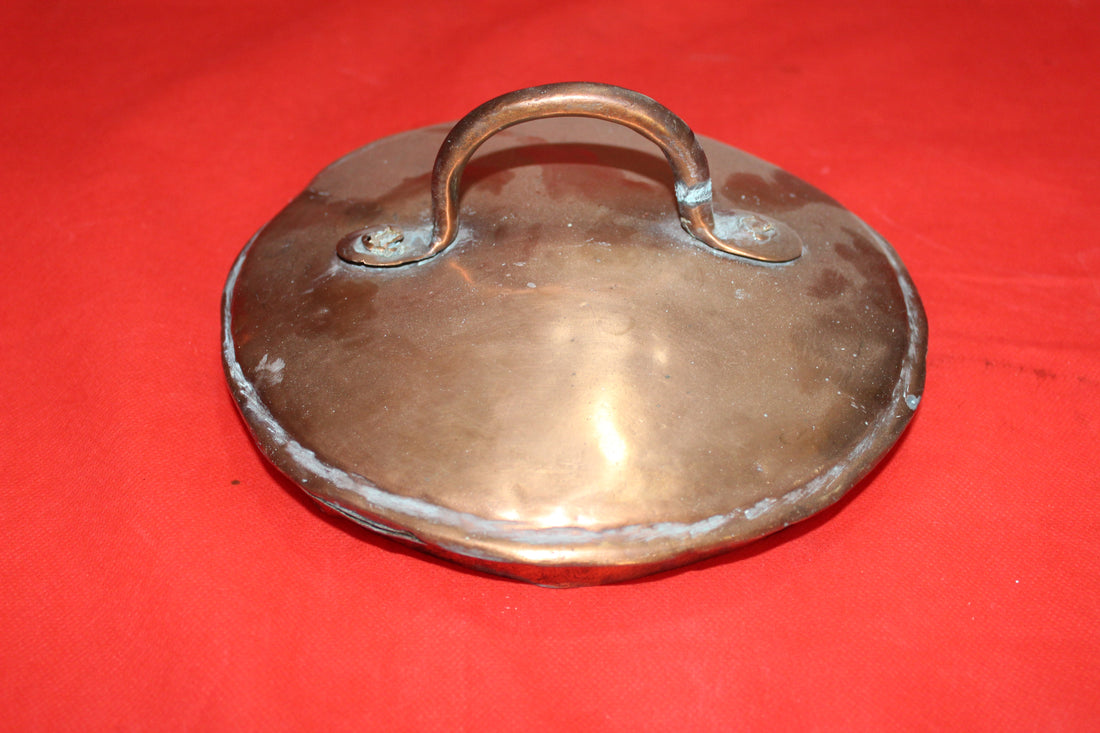 Tapadera de olla de cobre con acabado envejecido sobre fondo rojo, ideal para decoración rústica en cocina. Producto de excelente calidad.