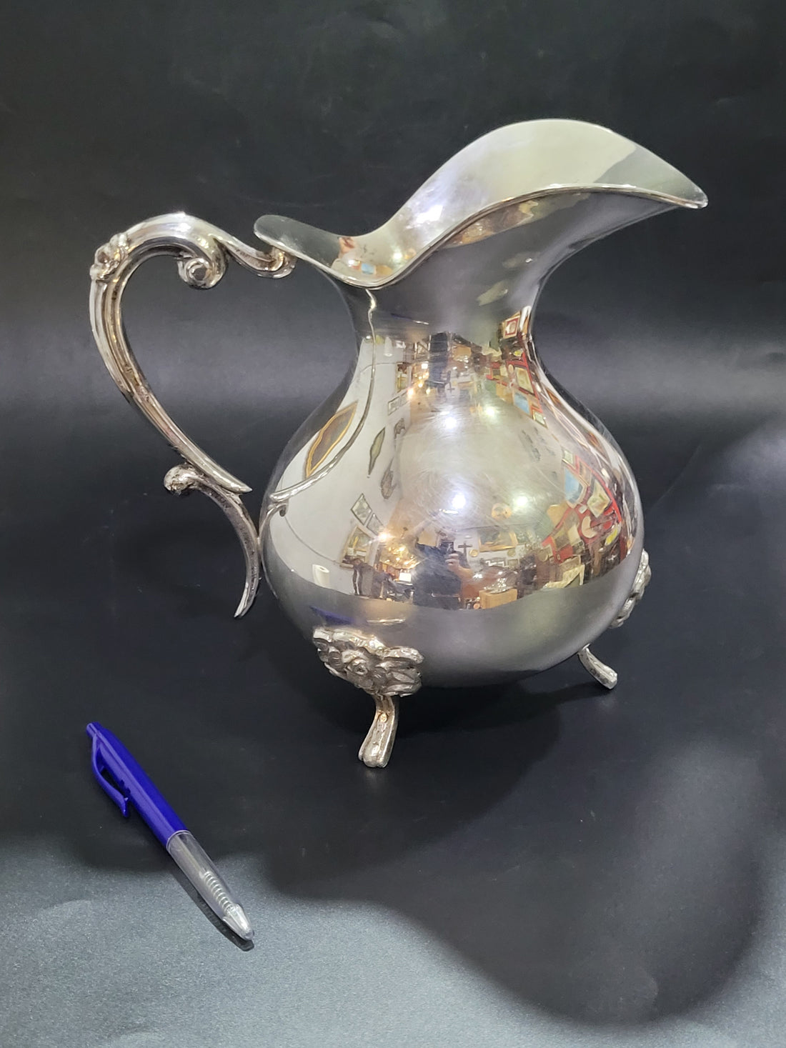 Vista lateral completa de la jarra de metal plateado sobre fondo negro, mostrando su asa ornamentada y su tamaño proporcional comparado con un bolígrafo.