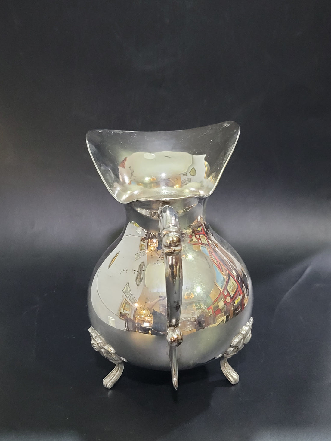 Vista lateral completa de la jarra de metal plateado sobre fondo negro, mostrando su asa ornamentada y su tamaño proporcional comparado con un bolígrafo.